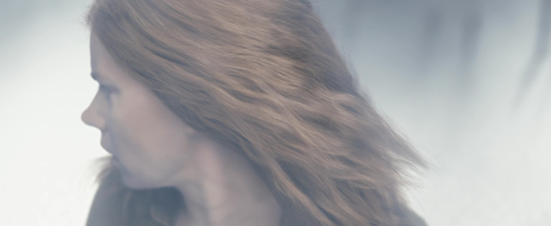 A red-haired woman in profile, turning away with her hair mid-motion against a hazy white background.